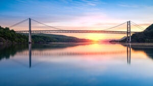 A wide bridge spanning a large, calm river, with the sun rising behind it